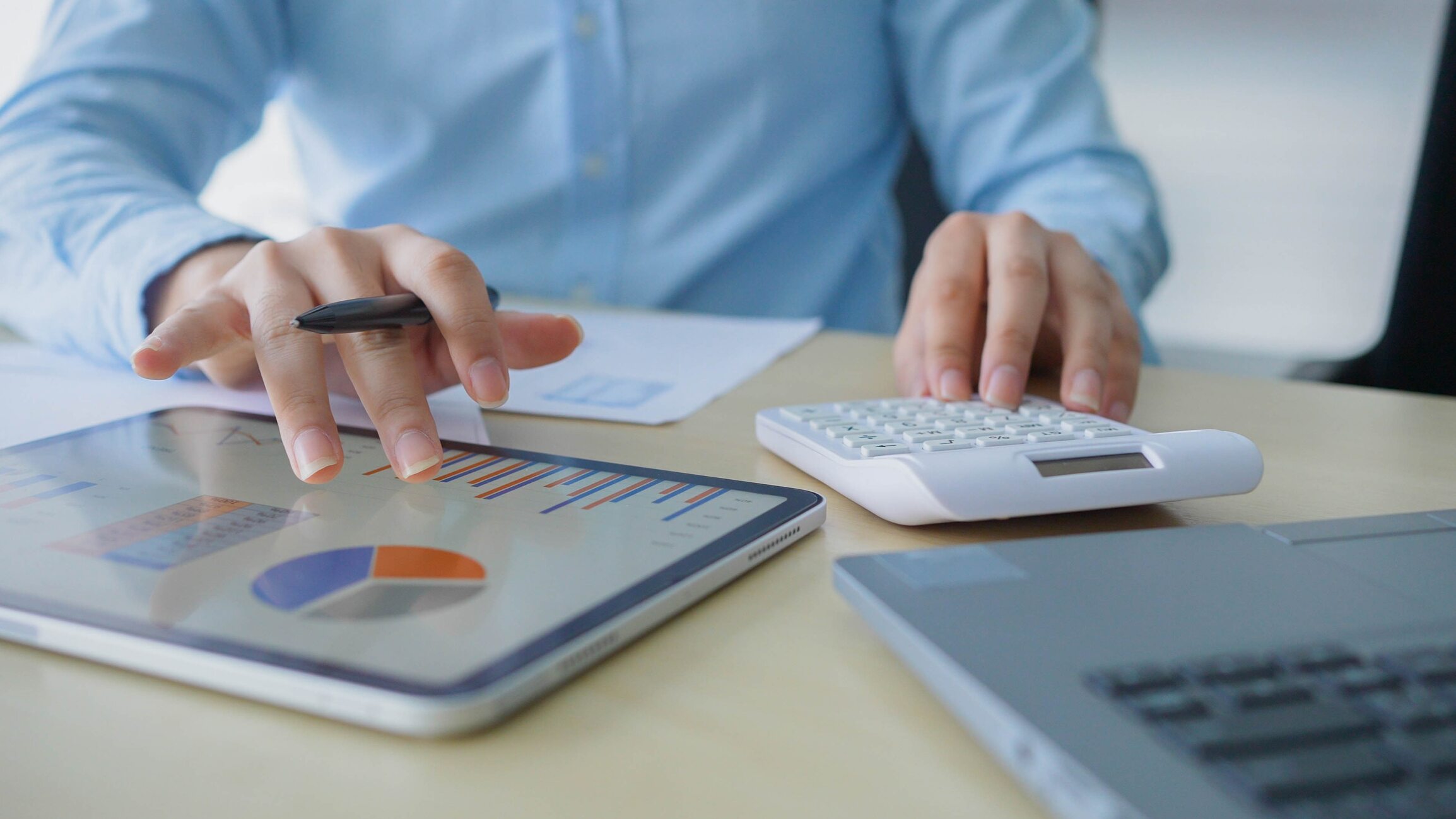 Man wearing a blue dress shirt uses a calculator and a tablet to examine data.